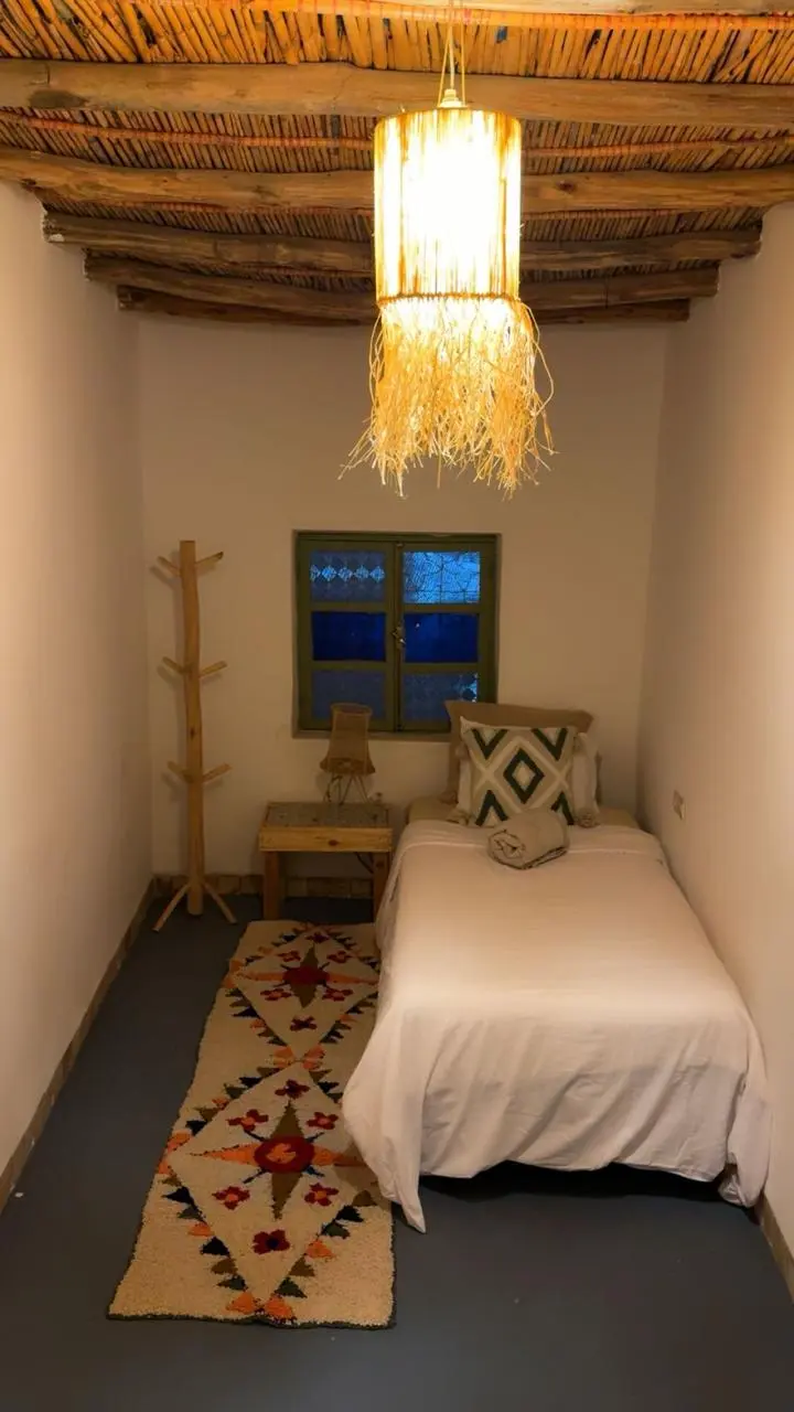 Chambre avec lit simple aux draps blancs, suspension en fibres naturelles, plafond en bois et tapis berbère